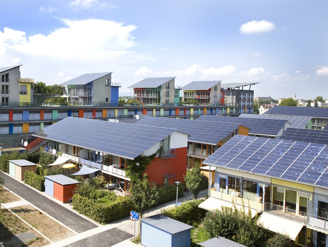 Luftaufnahme einer nachhaltigen Wohnsiedlung in Freiburg mit ausgedehnten Solaranlagen auf den Dächern. Die Gebäude zeigen farbenfrohe Fassaden und energieeffizientes Design, was Freiburgs Engagement für erneuerbare Energien und nachhaltige Stadtplanung demonstriert.